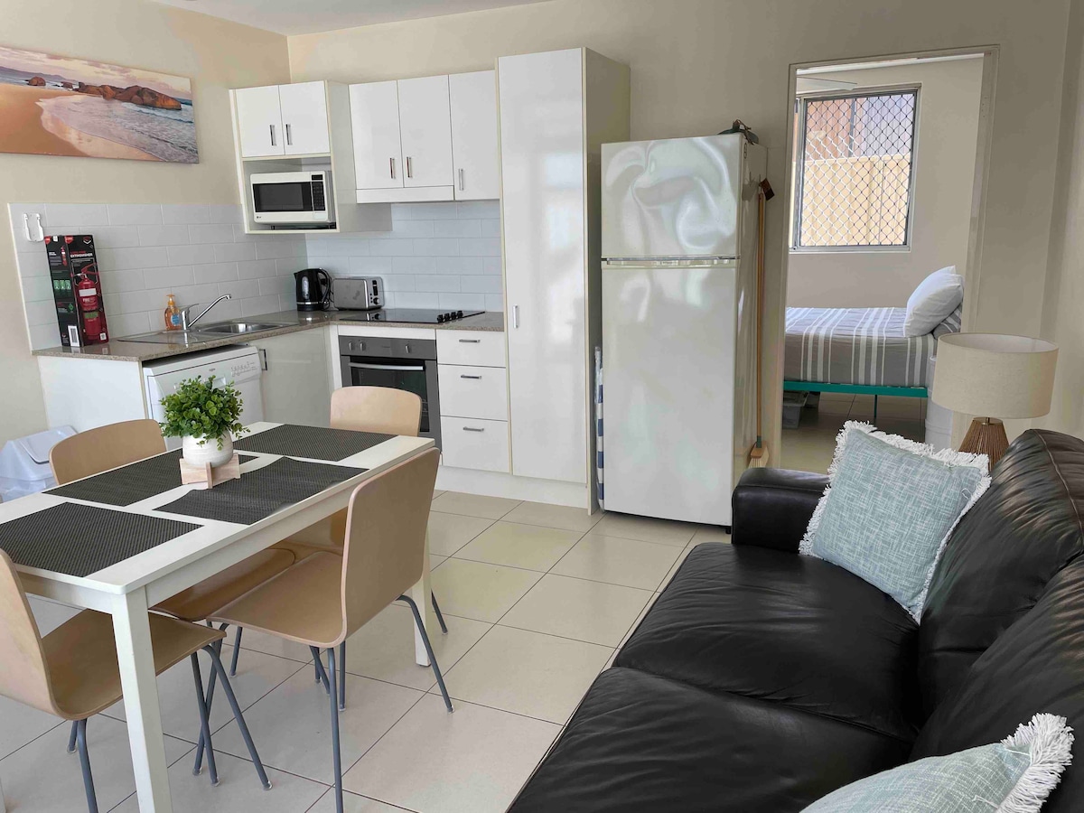 The open-plan living area features a dining table set for four, a well-equipped kitchen with modern appliances, and a comfortable lounge. A soft blue cushion adds a touch of color to the dark sofa. A separate bedroom with a bed is visible through the doorway.