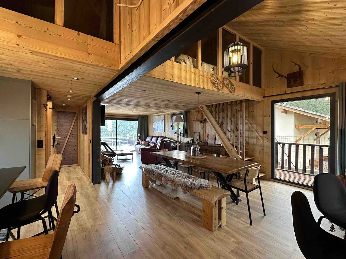 The spacious living area features warm wooden walls and a high ceiling with exposed beams. A large dining table is available, accompanied by a cozy seating area with a sofa. Large glass doors provide natural light and access to the outdoor terrace.