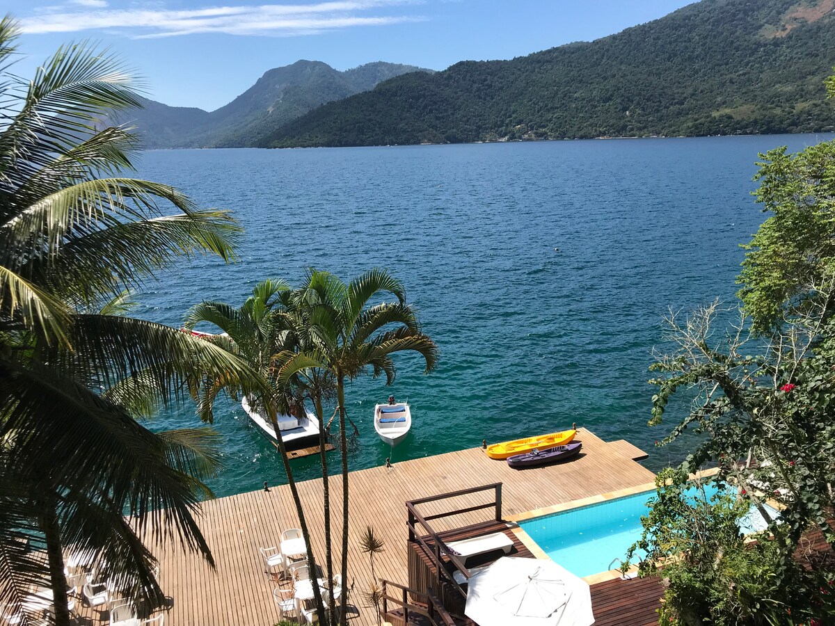 A scenic view of a wooden deck extending towards crystal-clear waters, with boats anchored nearby. Lush greenery surrounds the space, and a swimming pool reflects the sunlight against the backdrop of verdant hills and a clear blue sky.