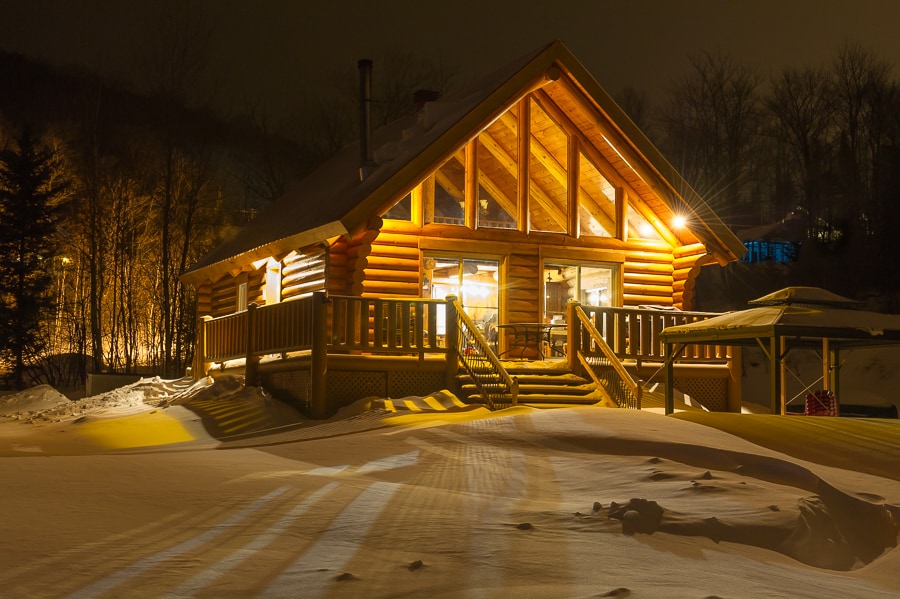 A log chalet is illuminated at night, showcasing a spacious deck leading to large glass windows. Snow blankets the surrounding landscape, while warm light emanates from inside, highlighting the wooden structure and sloped roof. Trees stand silhouetted in the background.