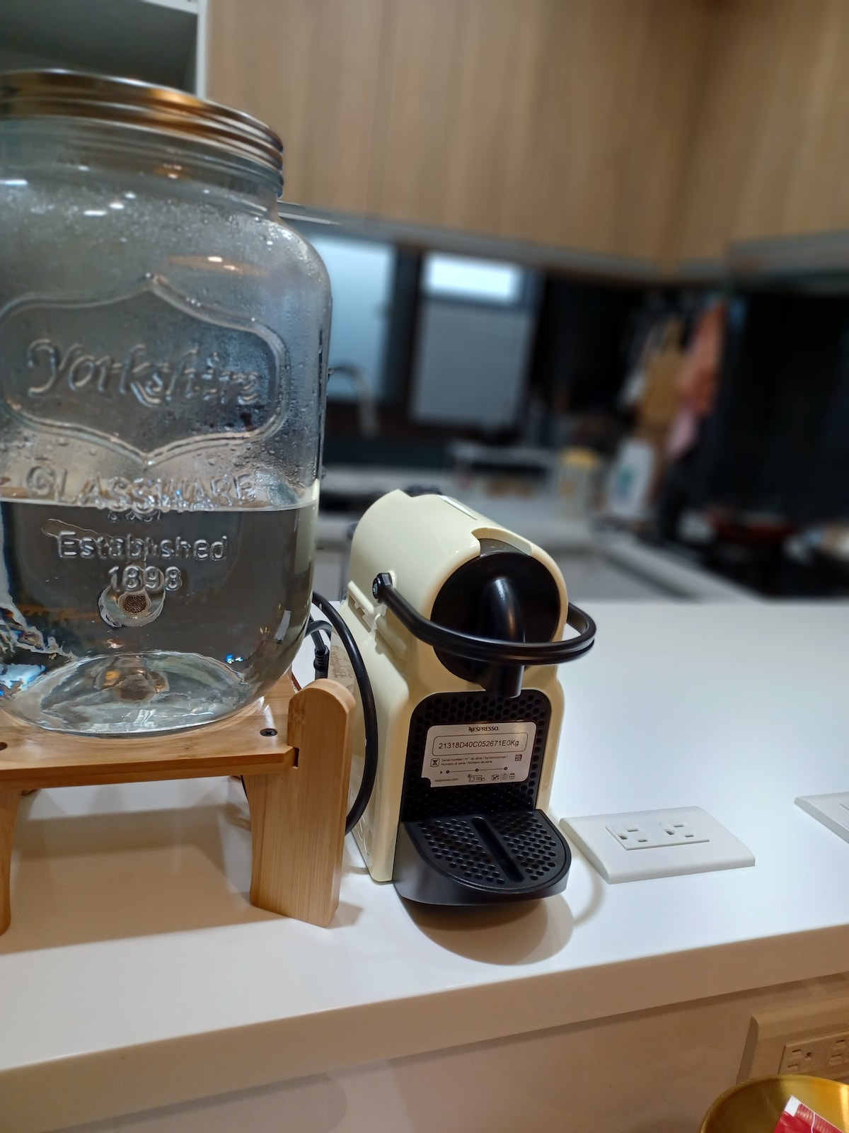 A sleek Nespresso capsule coffee machine is positioned on a clean countertop, next to a large, vintage-style glass jar filled with water. The background features wooden cabinetry and is subtly illuminated, creating a functional kitchen space.