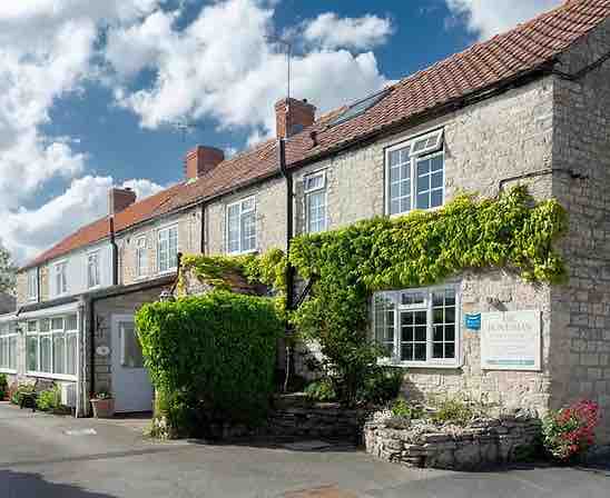 The exterior of Horseshoe Cottage features a traditional stone facade adorned with greenery. Bright windows and a welcoming entrance create a charming view. The cottage is set within a peaceful village, with a mix of historical and modern elements visible in the surrounding area.