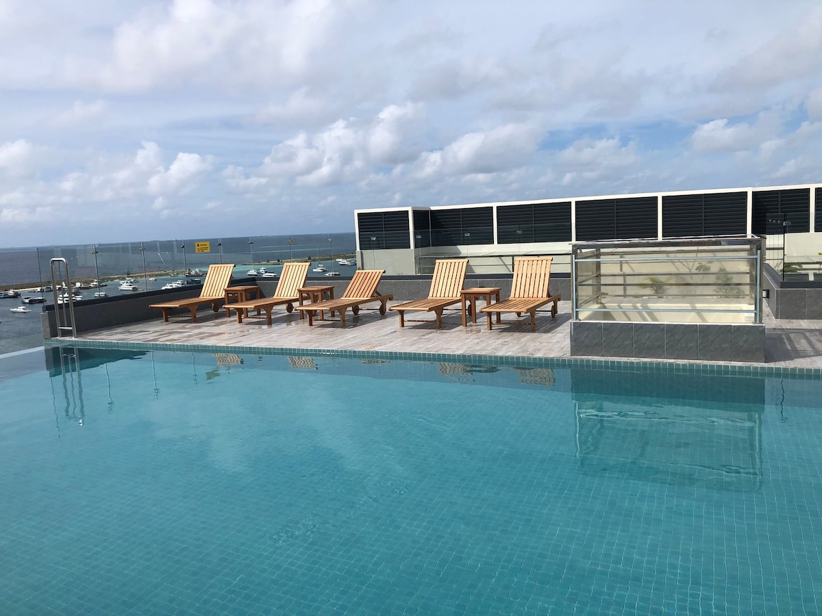 A rooftop swimming pool reflects the sky, surrounded by several wooden loungers positioned for relaxation. The area offers expansive views of the ocean and nearby boats, creating an inviting space for unwinding in a serene environment.