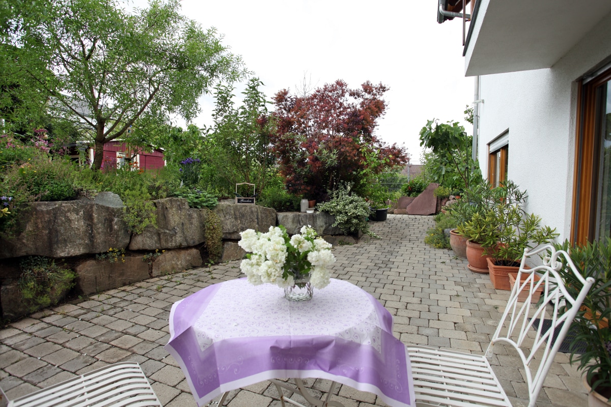 A charming outdoor space is presented, featuring a small table set with a vase of white flowers. Surrounding greenery and potted plants create a serene environment, while stone pathways invite relaxation. The area is flanked by a wall of natural stones and lush plants.