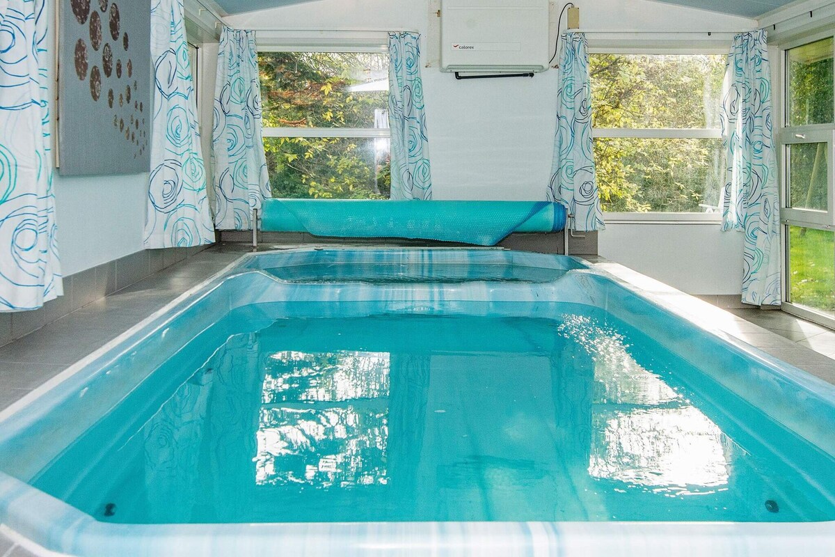 A spacious indoor pool area features a child-friendly pool with clear, inviting water. Large windows allow natural light to illuminate the space, while light-colored curtains add a soft contrast to the tiled flooring.
