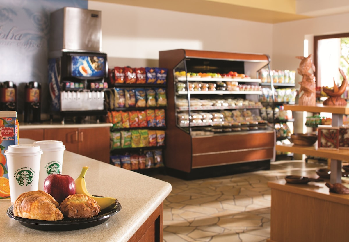 A well-lit convenience store is equipped with a variety of snacks and beverages. A display case showcases fresh meals and packaged items. A small table features two cups of coffee, a croissant, an apple, and a banana, inviting a quick dining experience.