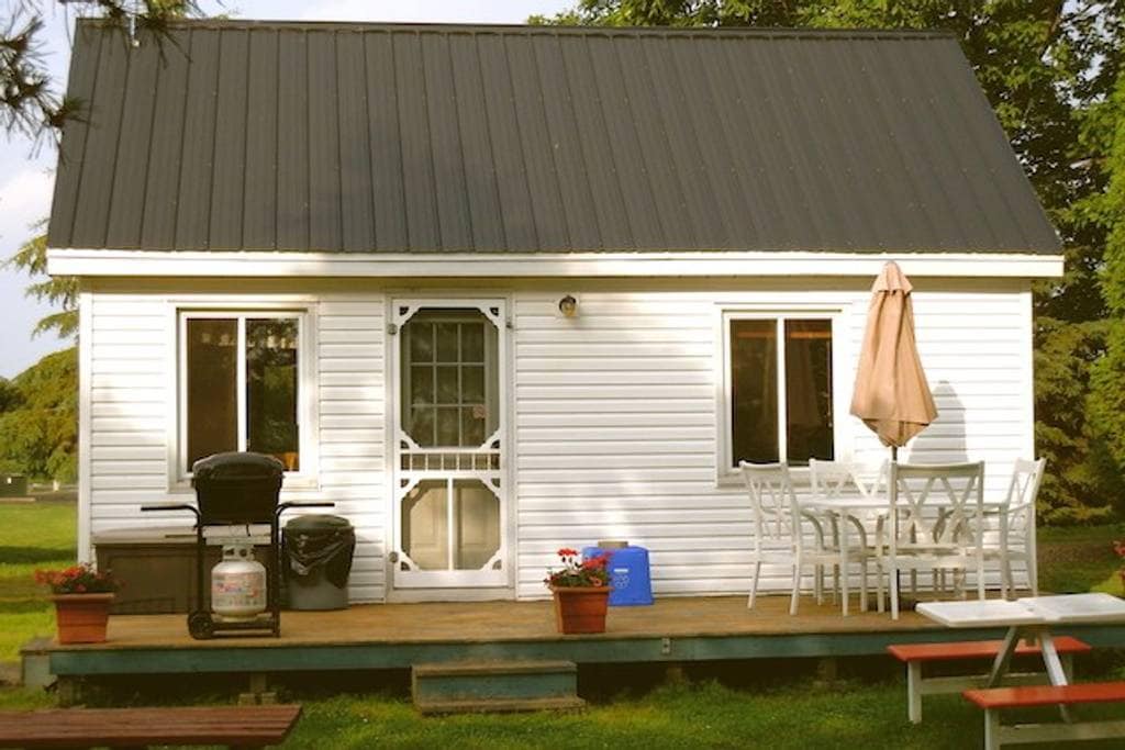 A white cottage is pictured, featuring a black metal roof. Two large windows are framed by flowering planters. A wooden deck is equipped with a table and chairs, as well as a barbecue grill, offering a space for outdoor relaxation.
