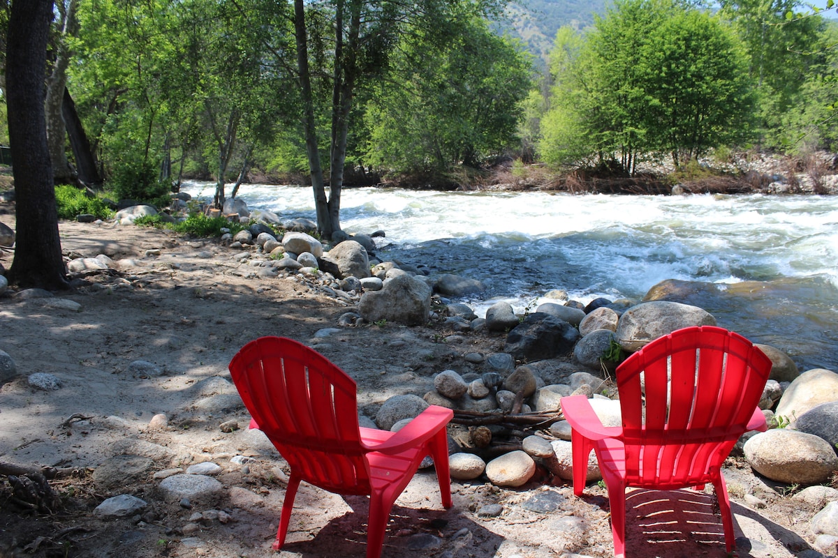 Two red Adirondack chairs are positioned by the riverbank, facing the flowing water. Stones outline a fire pit nearby, and lush greenery from surrounding trees adds a natural backdrop. The dynamic river’s movement is evident, creating a peaceful yet lively scene.