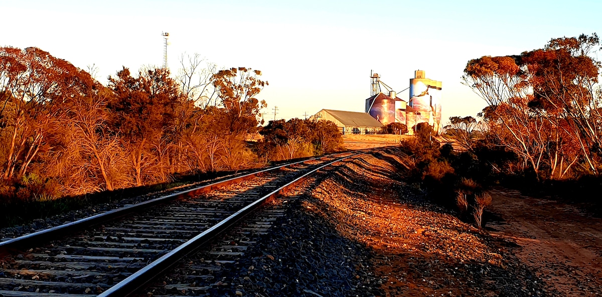 The image shows a railway track lined with gravel, leading toward industrial buildings in the background. The landscape is characterized by low vegetation with dried foliage, illuminated by the warm glow of sunset. Clear skies enhance the peaceful setting of the scene.