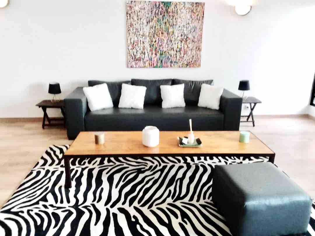 The living area features a dark sofa adorned with light-colored cushions, complemented by a wooden coffee table and an ottoman. A vibrant piece of wall art adds color, while a zebra-patterned rug enhances the space. Two small side tables with lamps are positioned beside the sofa.