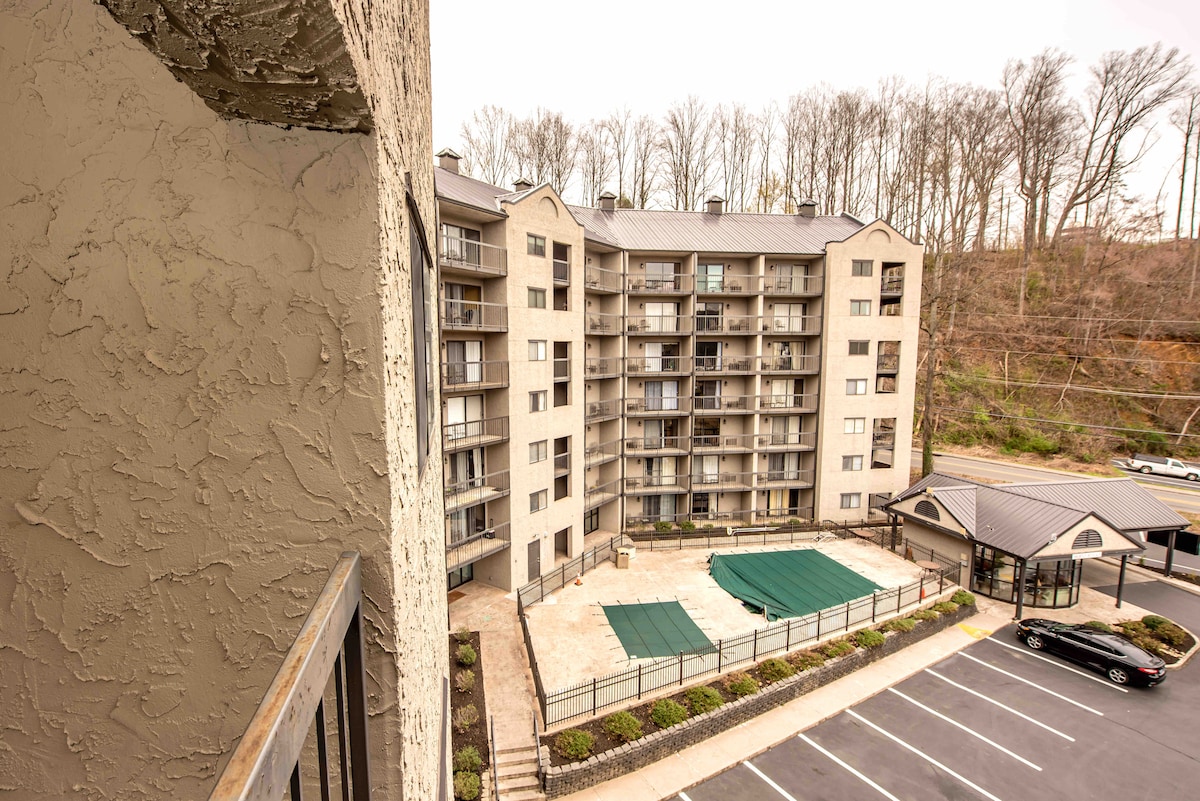 A view from the balcony overlooks the condo complex's outdoor area, featuring a pool with green covers and sun loungers. The building's exterior is composed of light-hued stucco, surrounded by trees, with a parking area visible in the foreground.