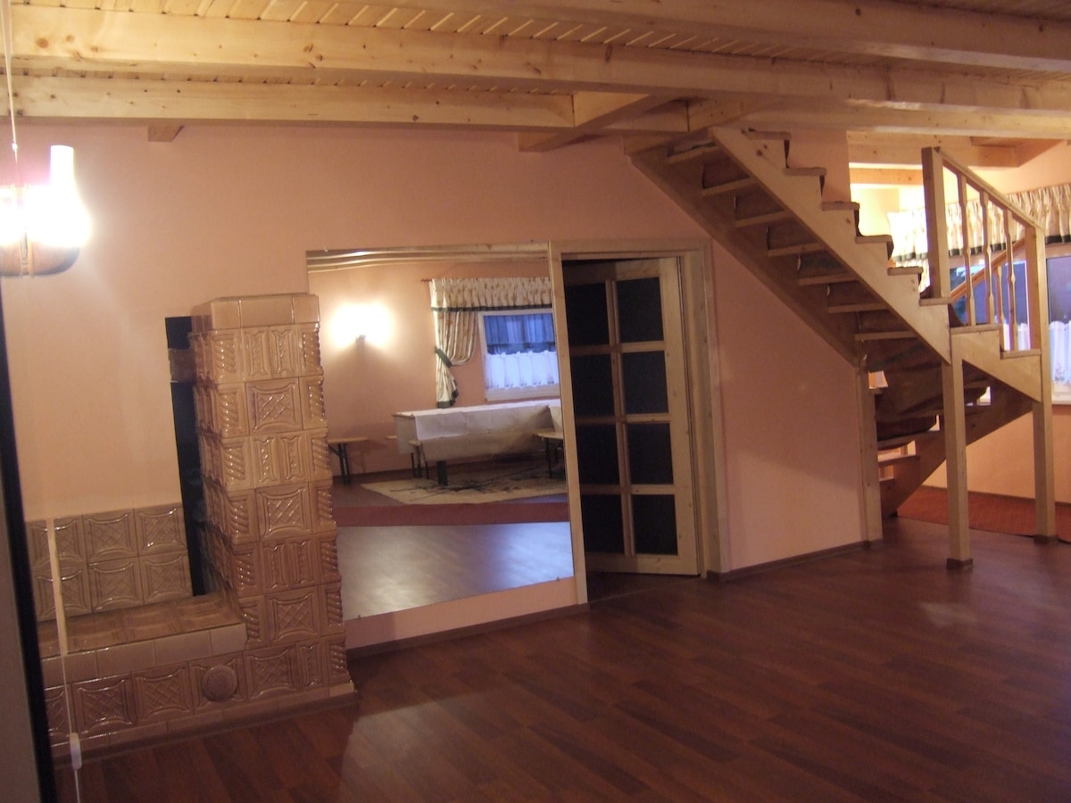An open living space features warm timber ceilings and wooden flooring. A staircase leads to the upper level. A large mirror reflects a dining area, while a traditional tile fireplace adds character. Soft lighting illuminates comfortable seating arrangements throughout the room.