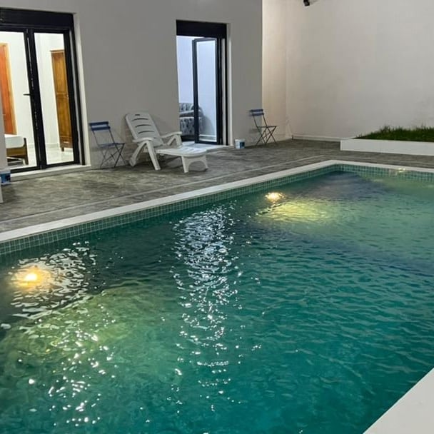 A clear swimming pool is visible, featuring gentle lighting that reflects off the water's surface. Two lounge chairs are placed adjacent to the pool on a spacious patio, while a grassy area is seen in the background. The entrance to the villa is located nearby.