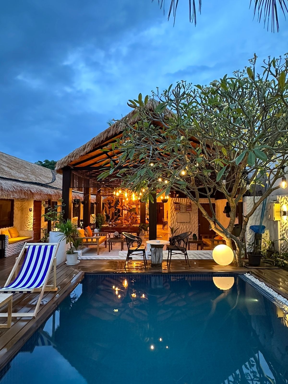 A serene outdoor area features a reflective pool beside a wooden deck adorned with tropical plants. Comfortable seating arrangements are illuminated under soft hanging lights, while a thatched roof provides shelter. The sky presents varying shades of blue as evening approaches.