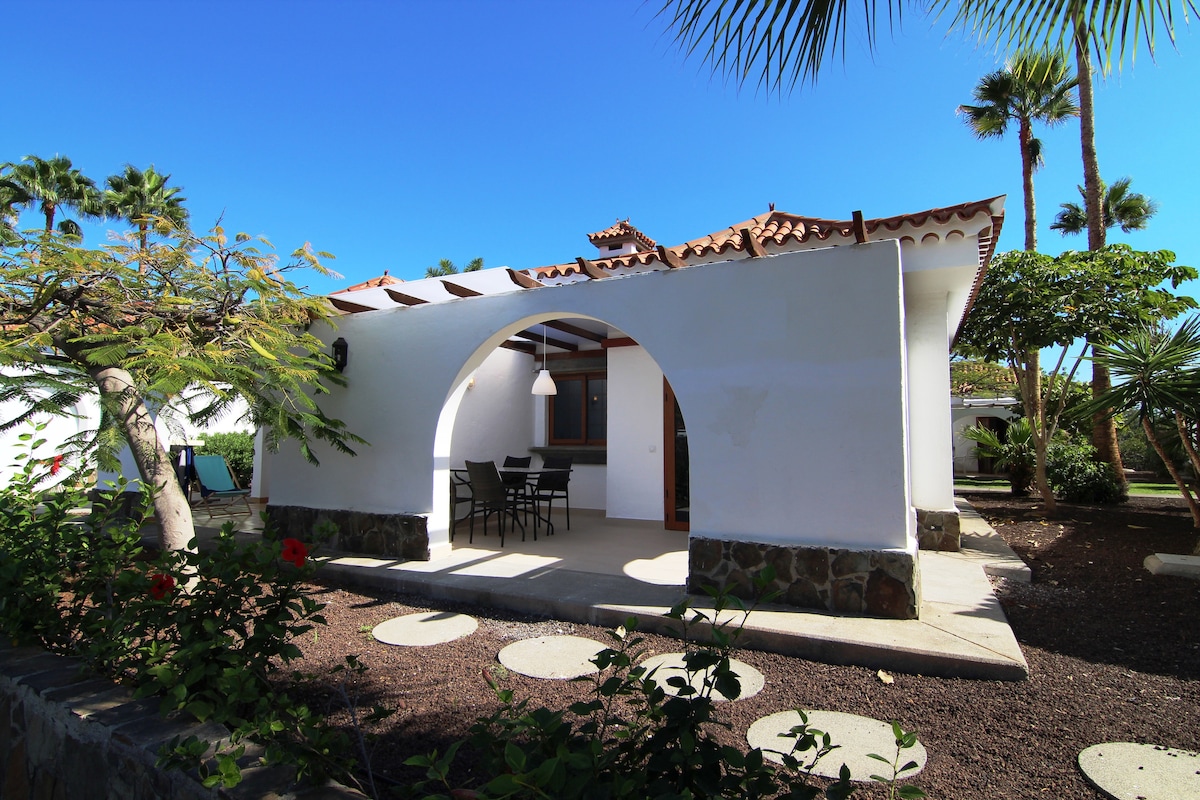 An exterior view of a bungalow is presented, featuring an arched entryway and a covered patio area. A dining set with chairs is positioned on the patio. Lush greenery and palm trees surround the space, enhancing the tranquility of the environment.