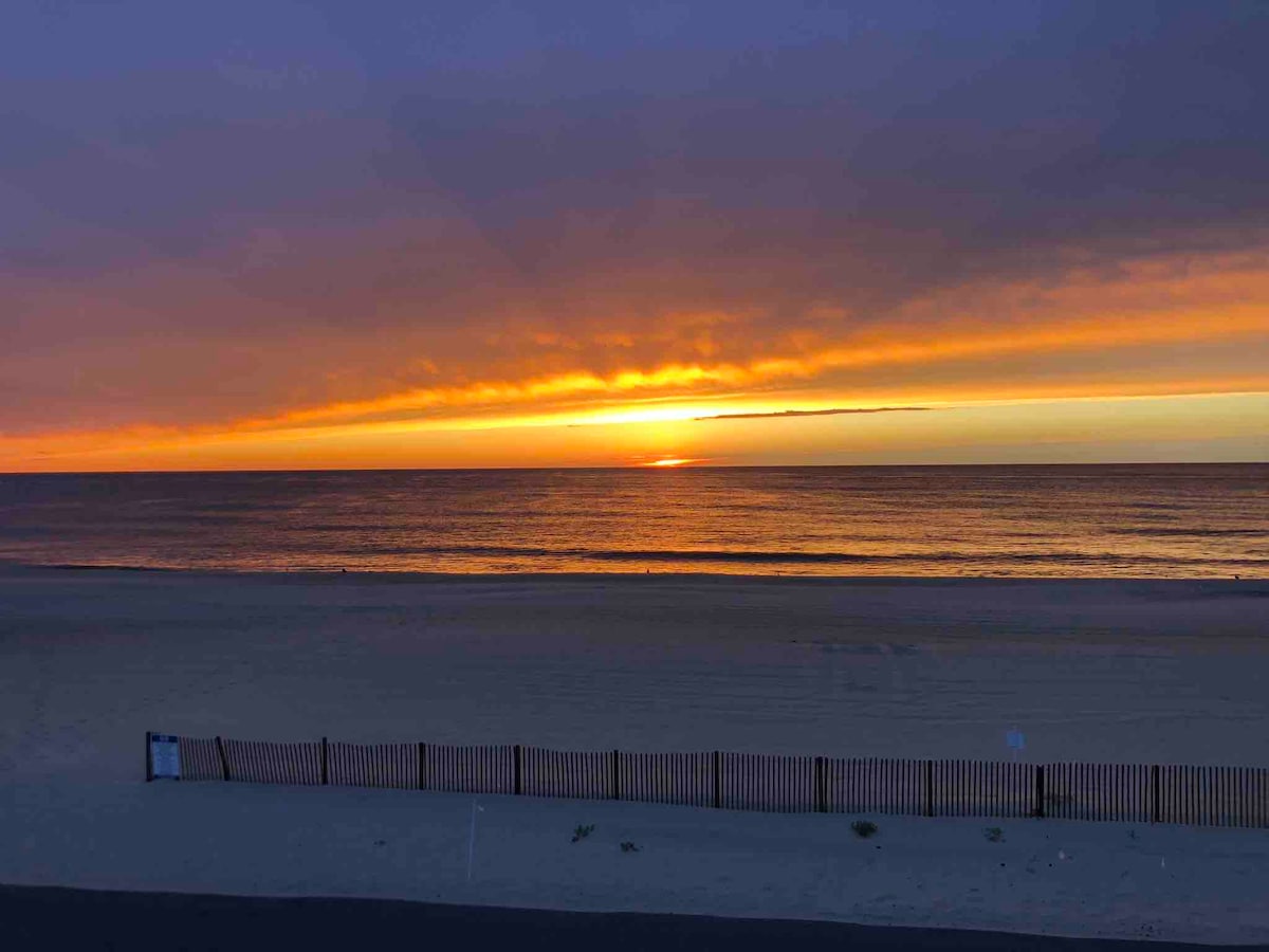 The image captures a serene sunrise over the ocean, displaying warm hues of orange and yellow blending into dark purple skies. The calm water reflects the vibrant colors, while a sandy beach stretches in the foreground, bordered by a wooden fence.