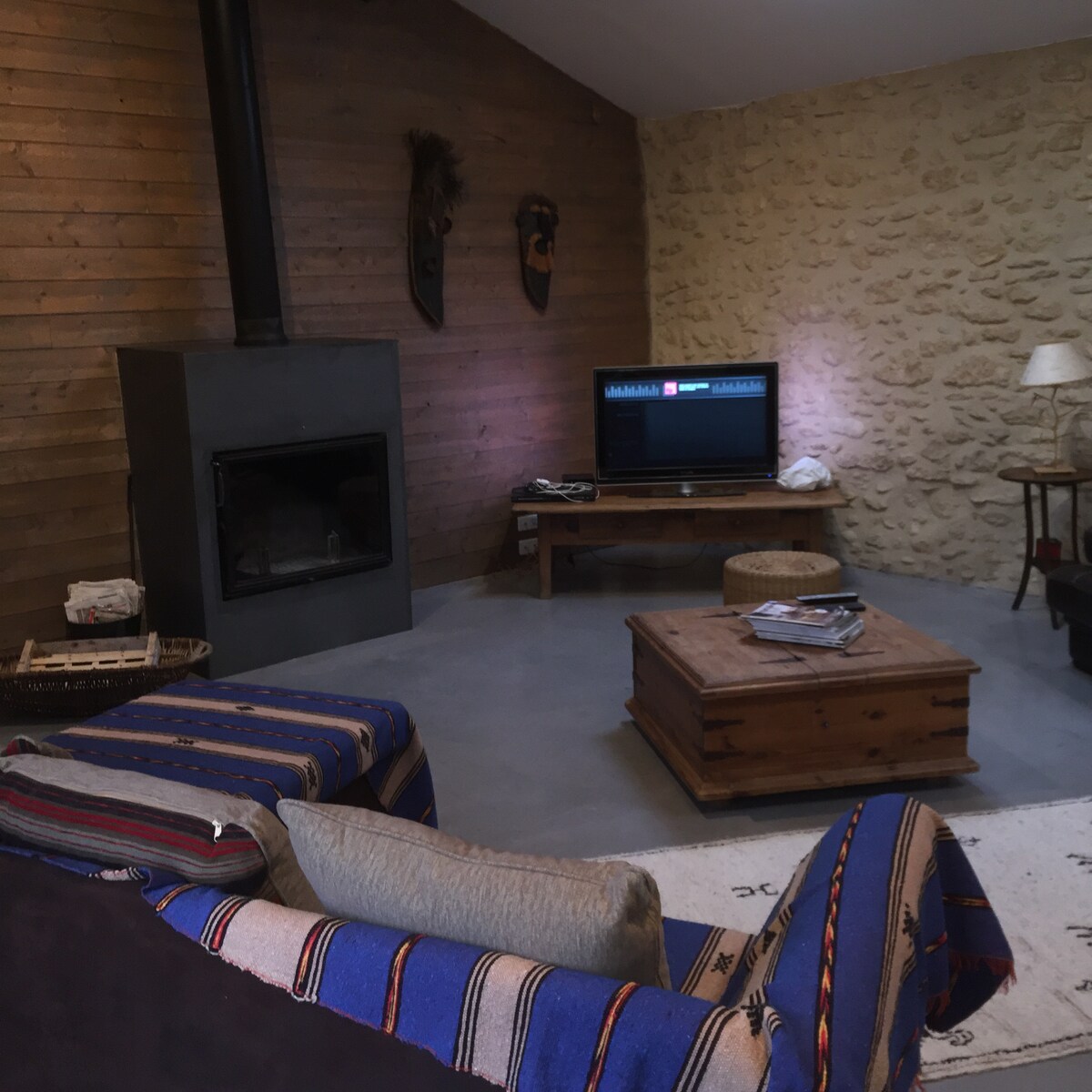 A cozy living area features a wood-burning stove against a textured stone wall. Comfortable seating is arranged around a wooden coffee table, with a television visible on a stand. Artistic wall masks add character to the space, enhancing its inviting ambiance.