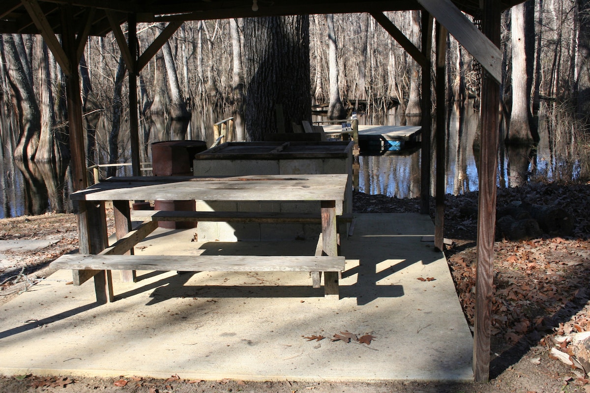 A shaded picnic shelter is featured, constructed with wooden beams and a concrete floor. A rectangular table and benches are provided, offering seating for guests. The calm water of the river is visible in the background, surrounded by trees reflecting in the water.