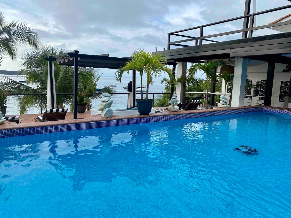 An outdoor pool with clear blue water occupies the foreground, surrounded by lush palm trees and comfortable lounge chairs. A deck area overlooks the ocean, with seating and shaded areas for relaxation. The sky is filled with soft clouds, enhancing the serene atmosphere.