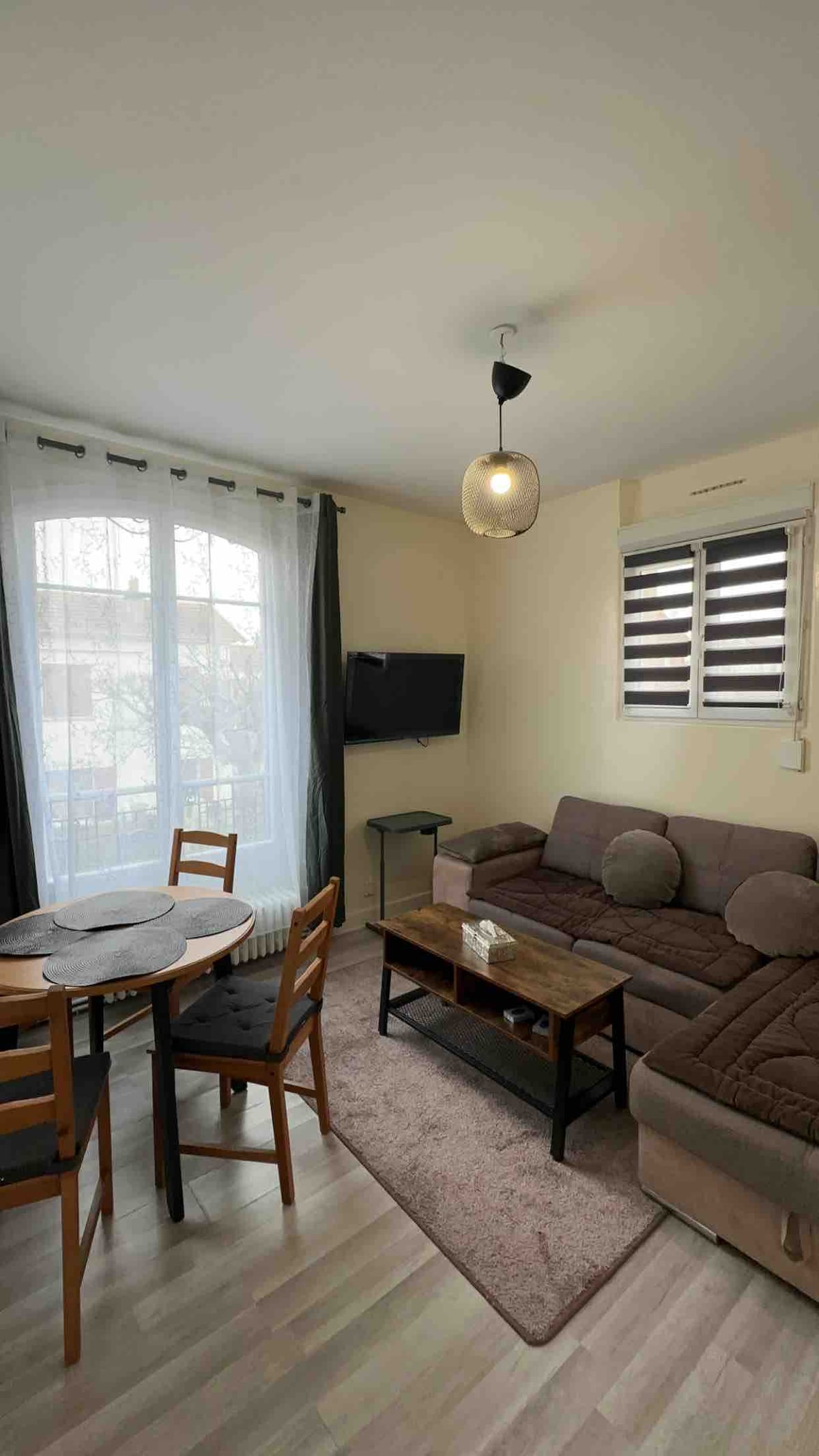 A cozy living area is presented with a comfortable sectional sofa and a wooden coffee table. A round dining table is paired with four wooden chairs. Natural light enters through large windows, adorned with light curtains, illuminating the neutral-toned space.