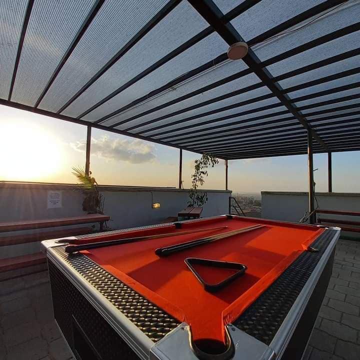 A rooftop chill spot features a pool table set under a retractable roof, providing shade and an outdoor experience. The sunset casts warm light on the space, while benches are arranged along the perimeter, offering a relaxed environment for socializing.