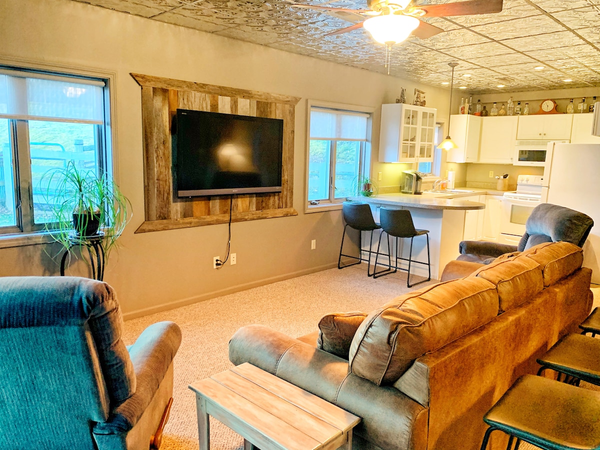 A comfortable living space is highlighted by a large flat-screen TV mounted on the wall. Two inviting recliner chairs and a spacious sofa provide seating, while a kitchen area features modern appliances and a breakfast bar with two stools. Natural light fills the room through multiple windows.