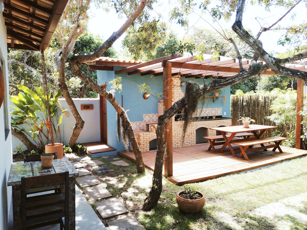 A serene outdoor space features a wooden deck with a large table and benches, complemented by a built-in barbecue area. Surrounding greenery includes leafy plants and trees, creating a tranquil environment. A stone pathway leads to the patio, enhancing accessibility and natural flow.