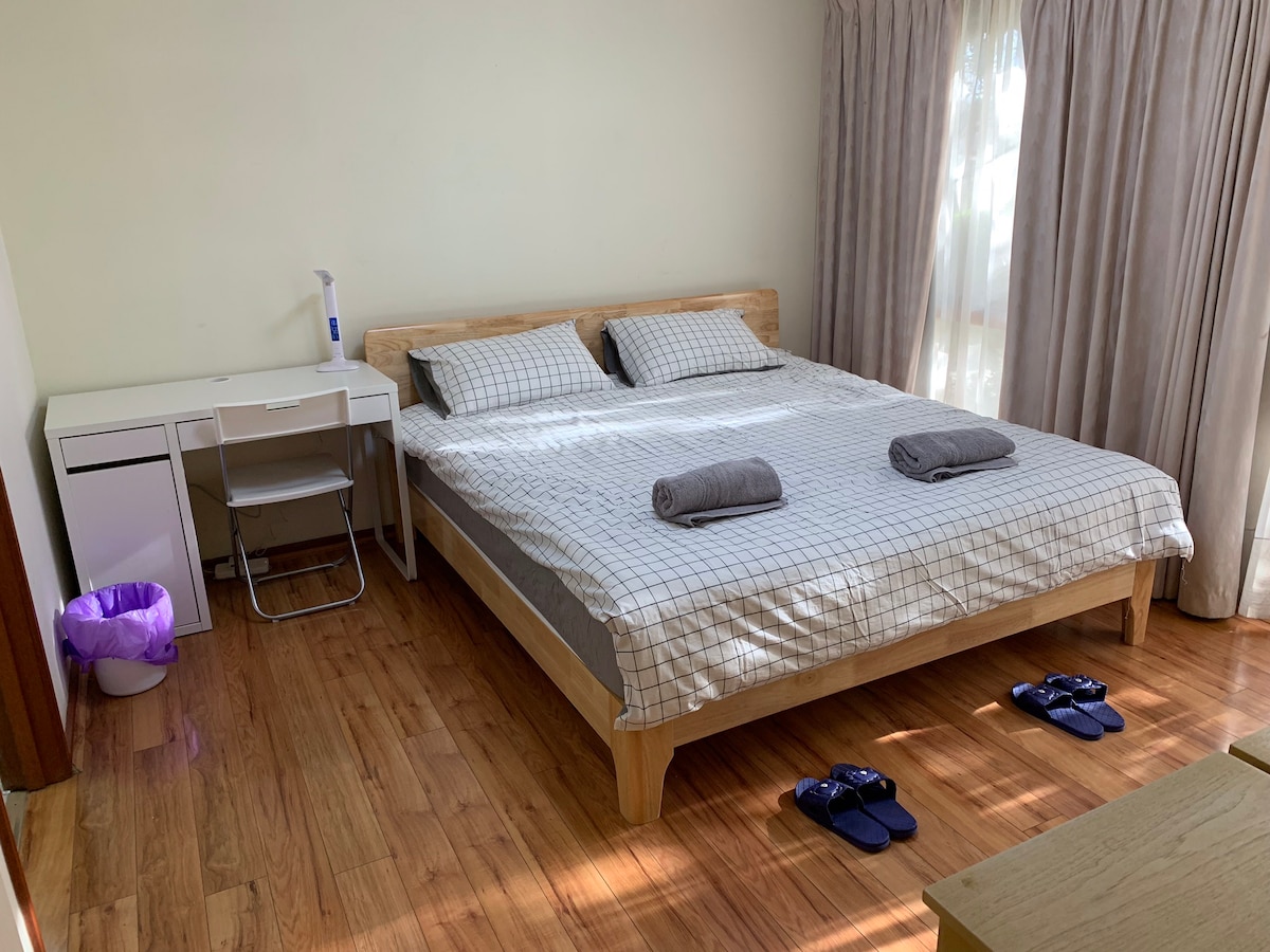 A spacious bedroom features a wooden bed with a light-colored checkered bedding arrangement. Two dark towels are neatly placed on the bed. A small desk and chair sit alongside a white bedside cabinet. Sunlight filters through sheer curtains, illuminating the warm-toned floor.