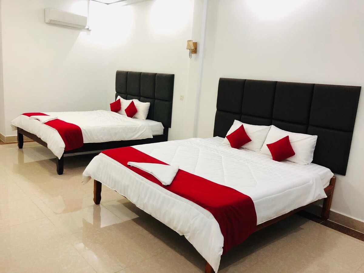 A bright room features two spacious beds, each adorned with red accents. The minimalist decor is complemented by neutral-colored walls, and natural light fills the space from above. Air conditioning is visible, and minimalist bedside lighting is mounted on the wall.