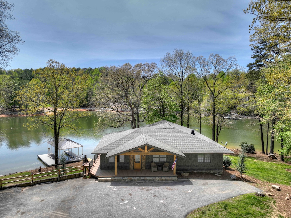 A cottage is situated close to the water's edge, surrounded by trees and greenery. A spacious deck extends from the front, offering views of the lake. The structure features a welcoming entrance and is complemented by a driveway leading to the entrance.