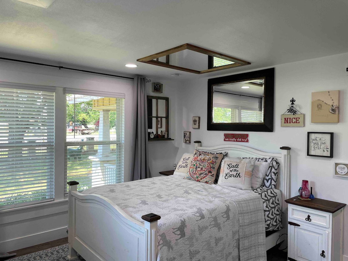A bright sleeping area features a spacious bed with decorative pillows and a light-colored quilt. Natural light enters through the window, which is dressed with sheer curtains. A mirror and a small shelf adorn the wall, complementing the room's cozy decor.