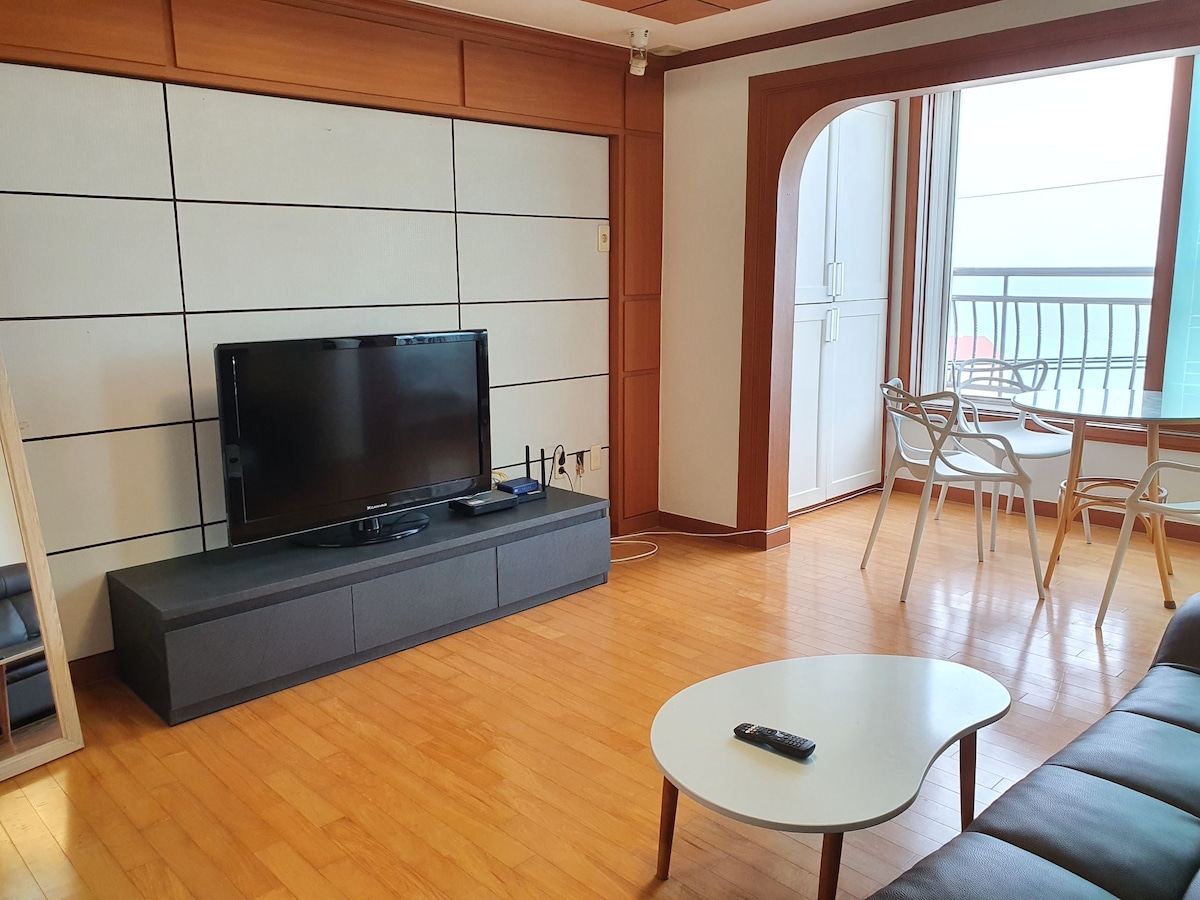A spacious living area features a modern television mounted on a low console against a backdrop of light wooden paneling. A round coffee table sits in front of a comfortable sofa. Nearby, a dining table with chairs provides a view through large windows.