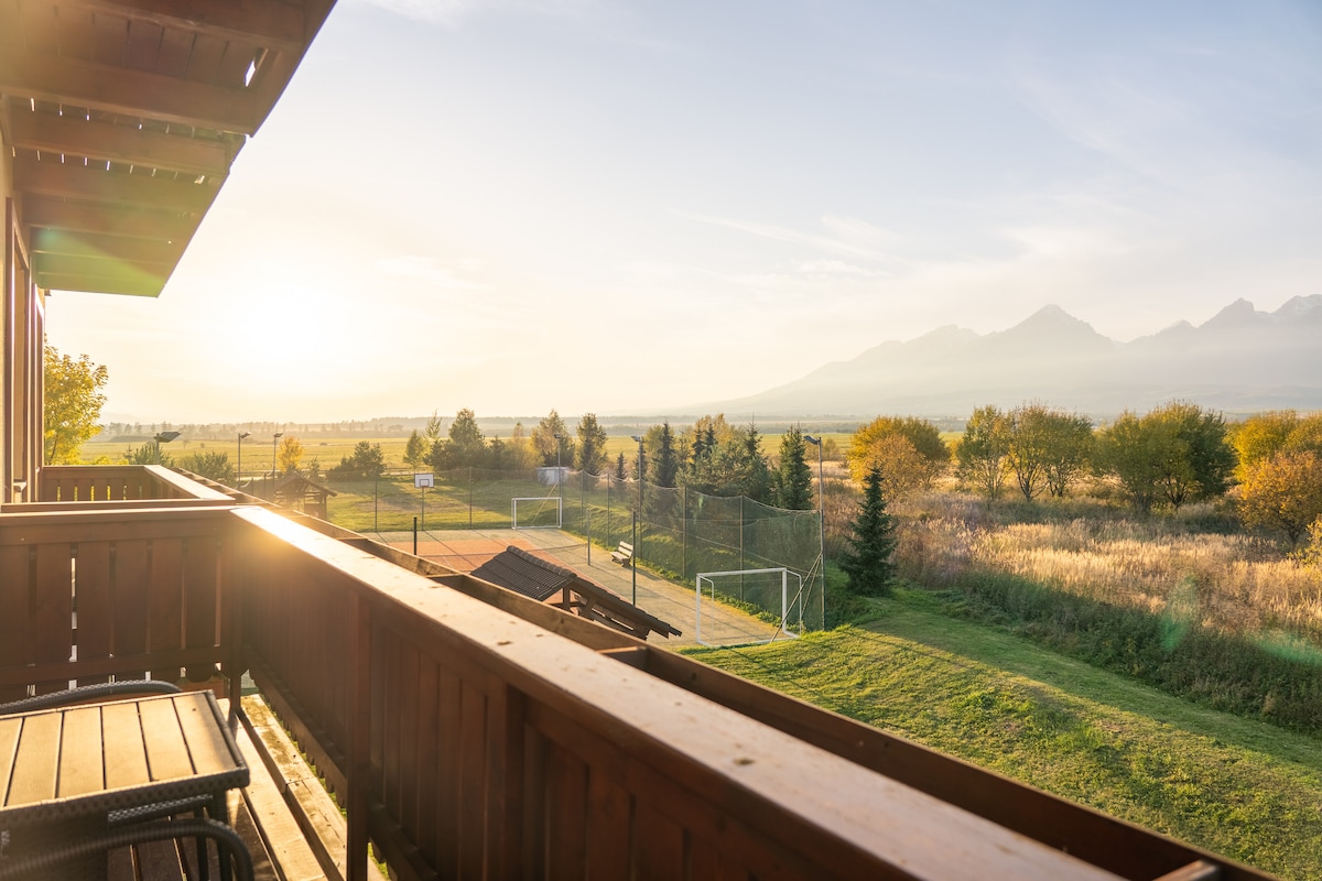A balcony view is presented, showcasing a wide expanse of green fields and sports courts below. The peaceful landscape is illuminated by soft light from the rising sun, with the distant mountains framed by a clear sky.