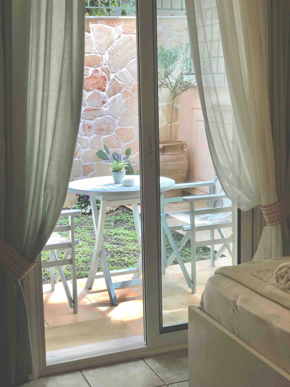 A cozy outdoor patio is visible through sliding glass doors, featuring a small round table and two chairs. Sunlight gently illuminates the space, complementing the greenery of the garden and decorative potted plants in the background.