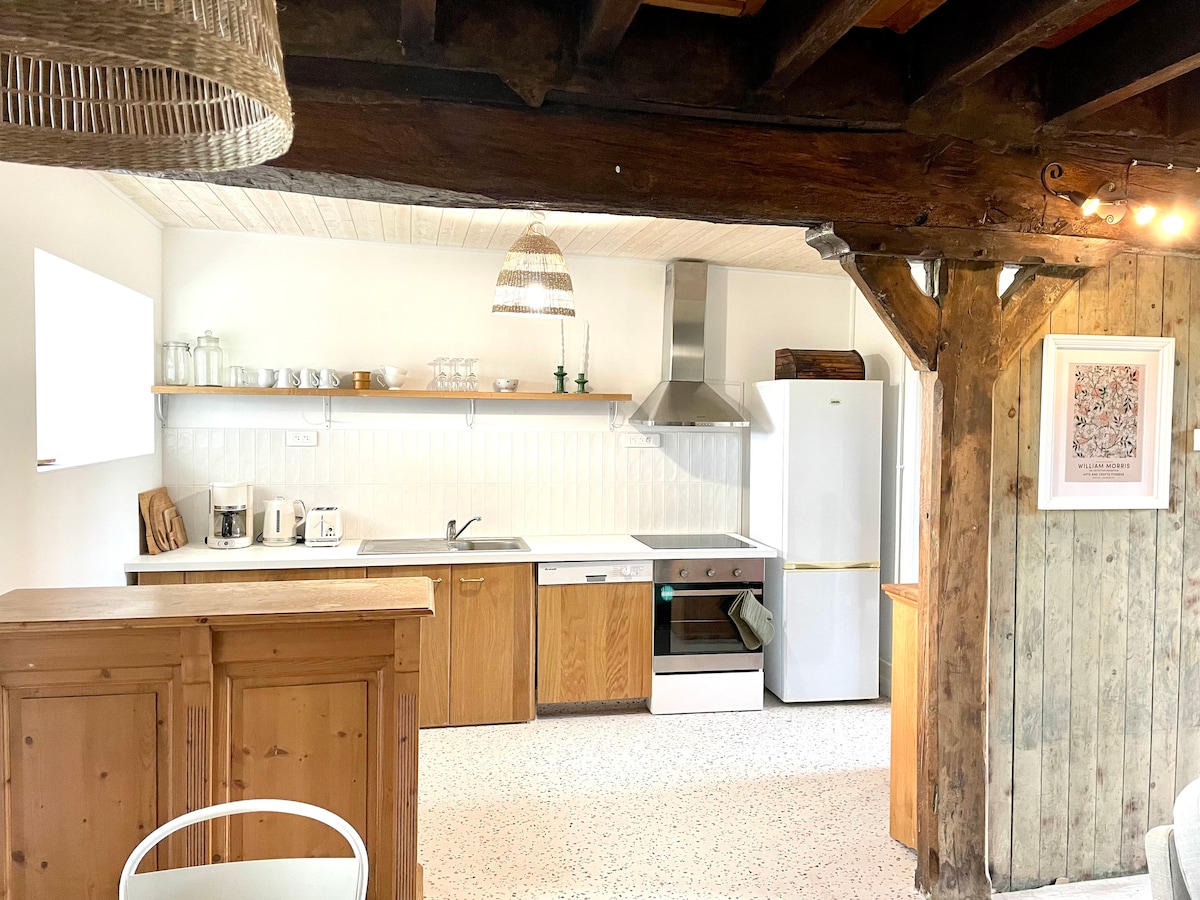 An open kitchen is featured with wooden cabinetry and modern appliances, including a refrigerator and oven. A spacious counter is accentuated by rustic beams, while natural light flows through a nearby window. Decorative elements, like a woven lamp, enhance the inviting atmosphere.
