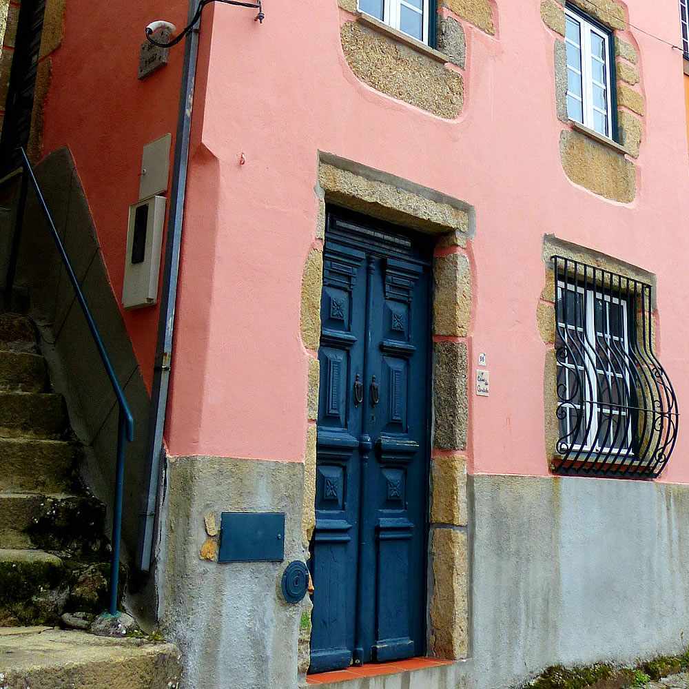 The exterior shows a charming pink building with textured stone accents. A dark blue door, featuring intricate designs, is set within a stone frame. Two small windows, secured with black bars, offer glimpses of the interior. Steps lead up to the entrance.