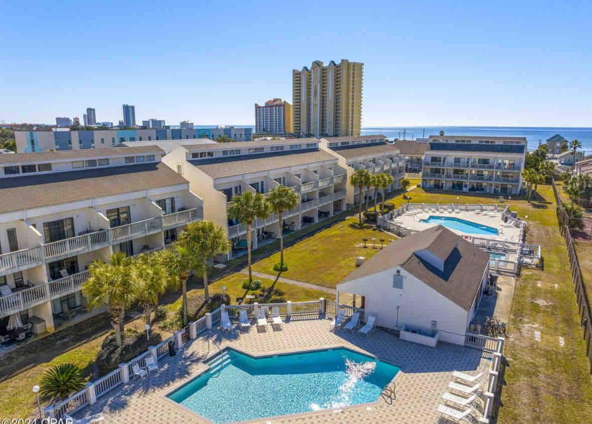 The image captures the serene courtyard of the condo complex, featuring two sparkling pools surrounded by lounge chairs. Lush palm trees provide greenery, and the beach is visible in the background, enhancing the tranquil setting. The buildings offer comfortable accommodations for guests.