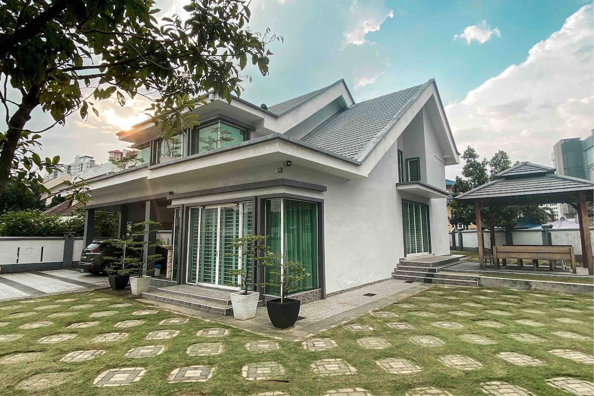 A modern two-story villa is presented, featuring large glass windows that reflect sunlight. The exterior showcases a manicured lawn with geometric paving stones, and a shaded gazebo is visible in the background, enhancing the outdoor space.