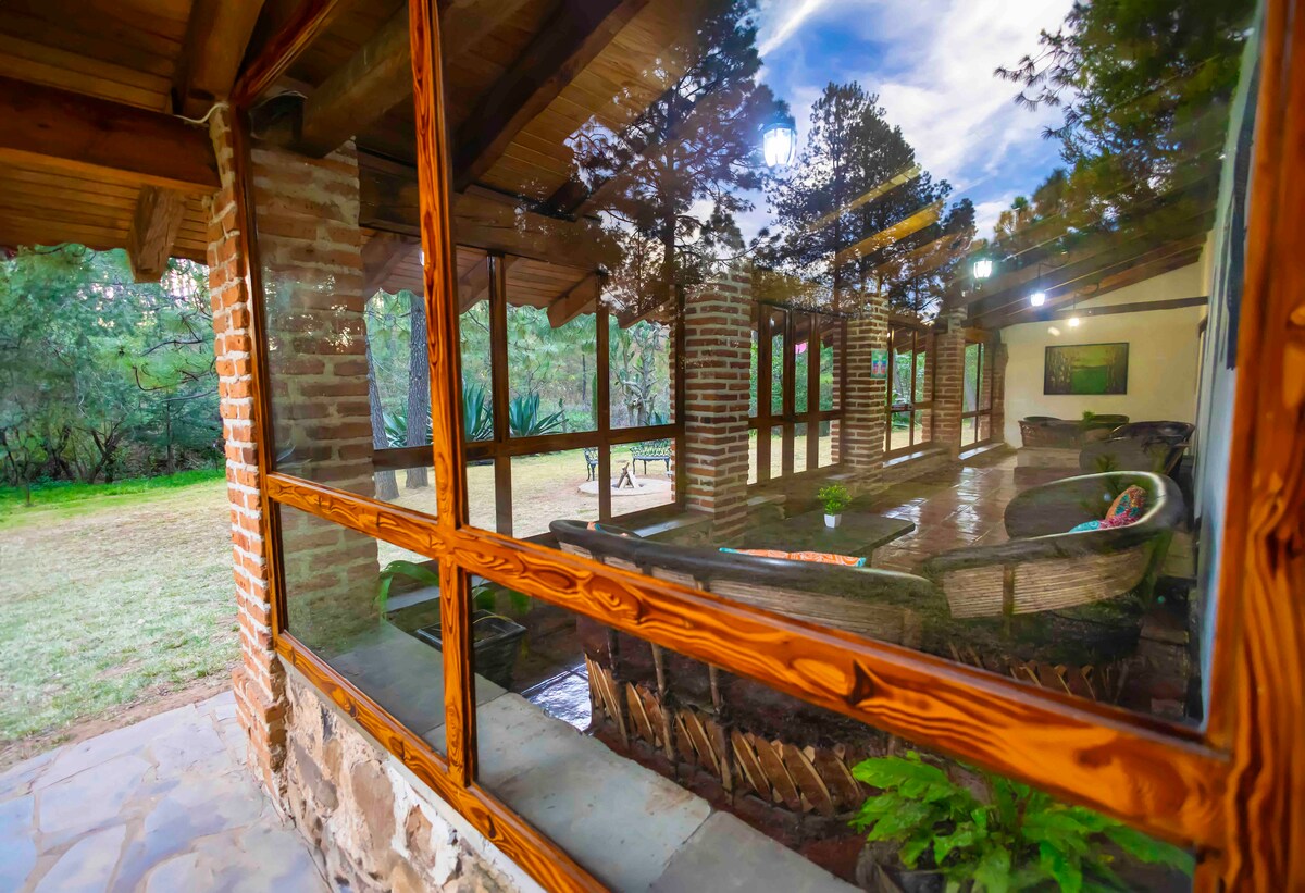A spacious glass-enclosed terrace offers views of the surrounding forest, enhancing the connection with nature. Wooden beams and brick columns provide structural interest, while the polished stone floor reflects the ambient lighting. Comfortable seating areas can be seen, inviting relaxation throughout the year.