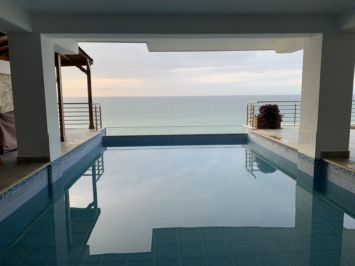 An expansive swimming pool is featured, with clear blue water reflecting the calm ocean view. The surrounding area includes a covered patio that provides shade, while the horizon showcases subtle tones of sky meeting water, enhancing the serene ambiance of the space.