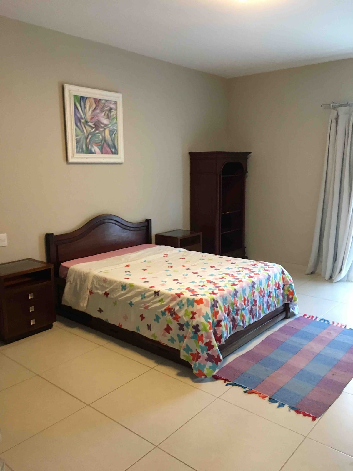 A cozy bedroom is highlighted by a decorated bedspread featuring colorful patterns. A wooden nightstand is positioned beside the bed, and a matching wooden cabinet adds function. Light-colored walls complement the natural light from a window dressed with a sheer curtain.