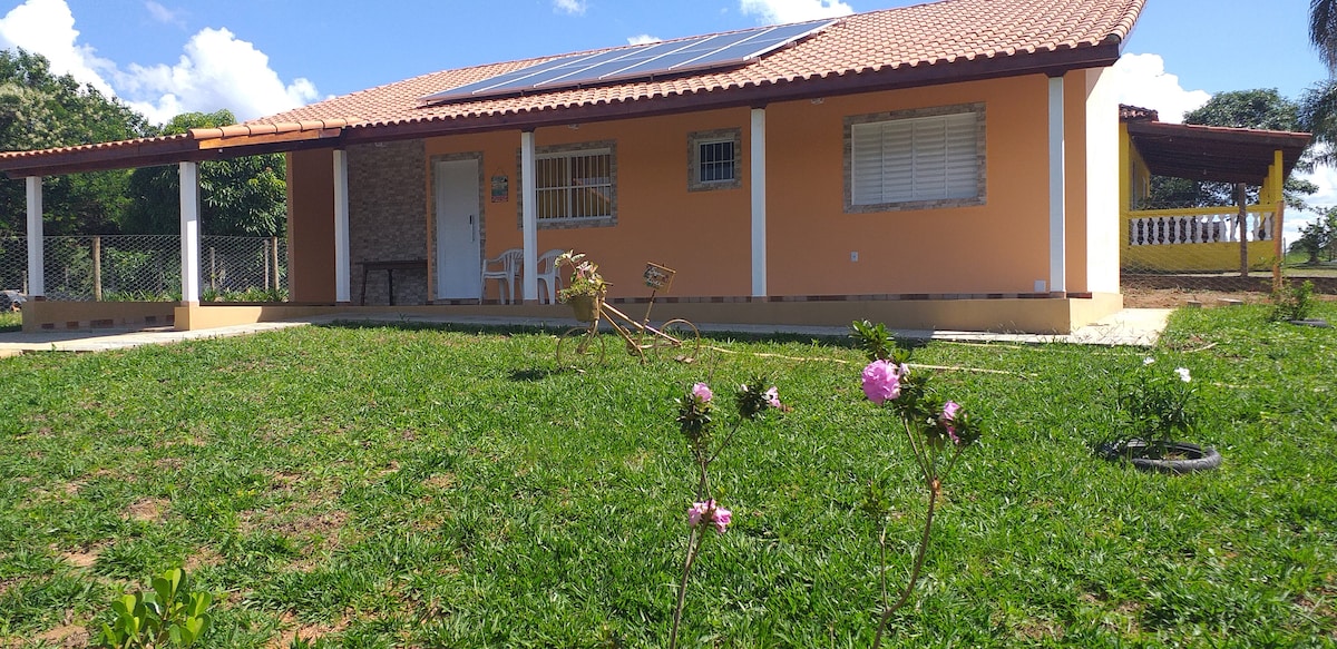The house features a bright exterior with a complementary roof, set against a lush green lawn. Solar panels are visible on the roof, and small flowering plants are planted in the foreground. A porch provides shaded outdoor space, enhancing the surrounding natural environment.