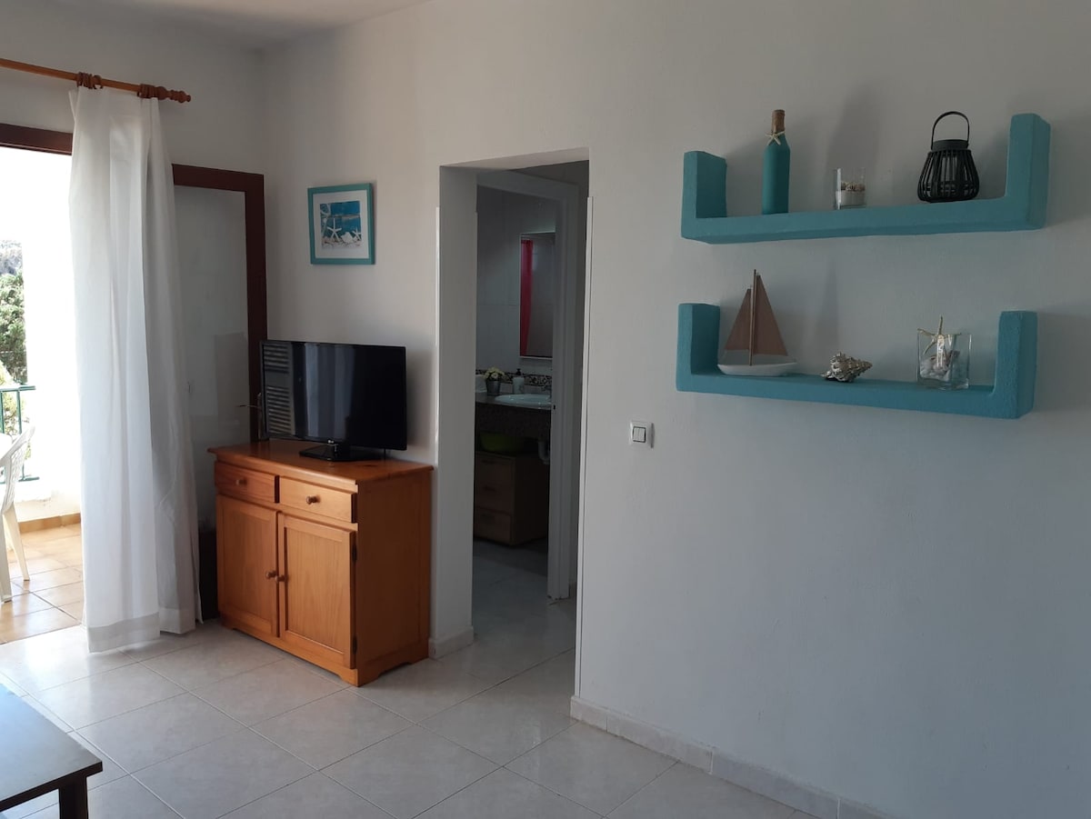 A light-filled living area features a wooden sideboard with a television and decorative items on a teal shelf. An open doorway leads to a kitchen area, with a white curtain softly filtering natural light. The tiled floor contributes to the airy ambiance of the space.