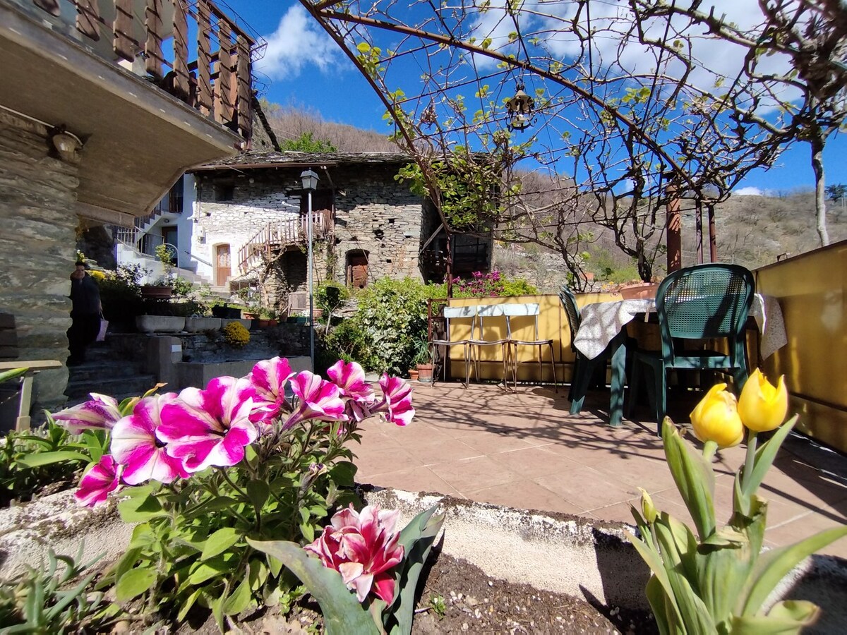 A charming garden area is surrounded by blooming flowers and greenery. A shaded patio features a dining table and chairs, offering space for relaxation. Stone walls and rustic buildings are visible in the background, enhancing the natural ambiance of the outdoor setting.