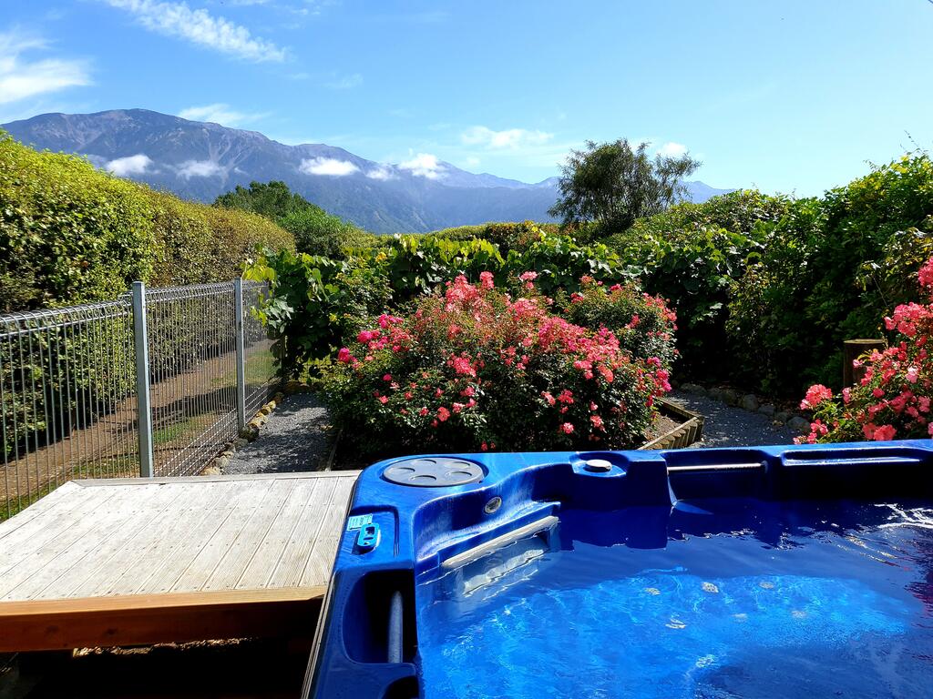 A hot tub with crystal-clear water is set against a backdrop of lush greenery and vibrant flowers. Scenic mountain views are visible in the distance, enhancing the serene outdoor environment. Wooden decking surrounds the hot tub, providing a relaxing space amid nature.