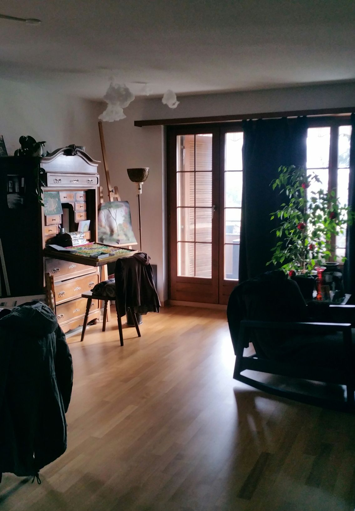 A bright corner of a living space features a wooden desk adorned with art supplies and an easel. Natural light comes through double glass doors with wooden frames, while a potted plant adds greenery to the area. A cozy chair and draped garments complete the setting.