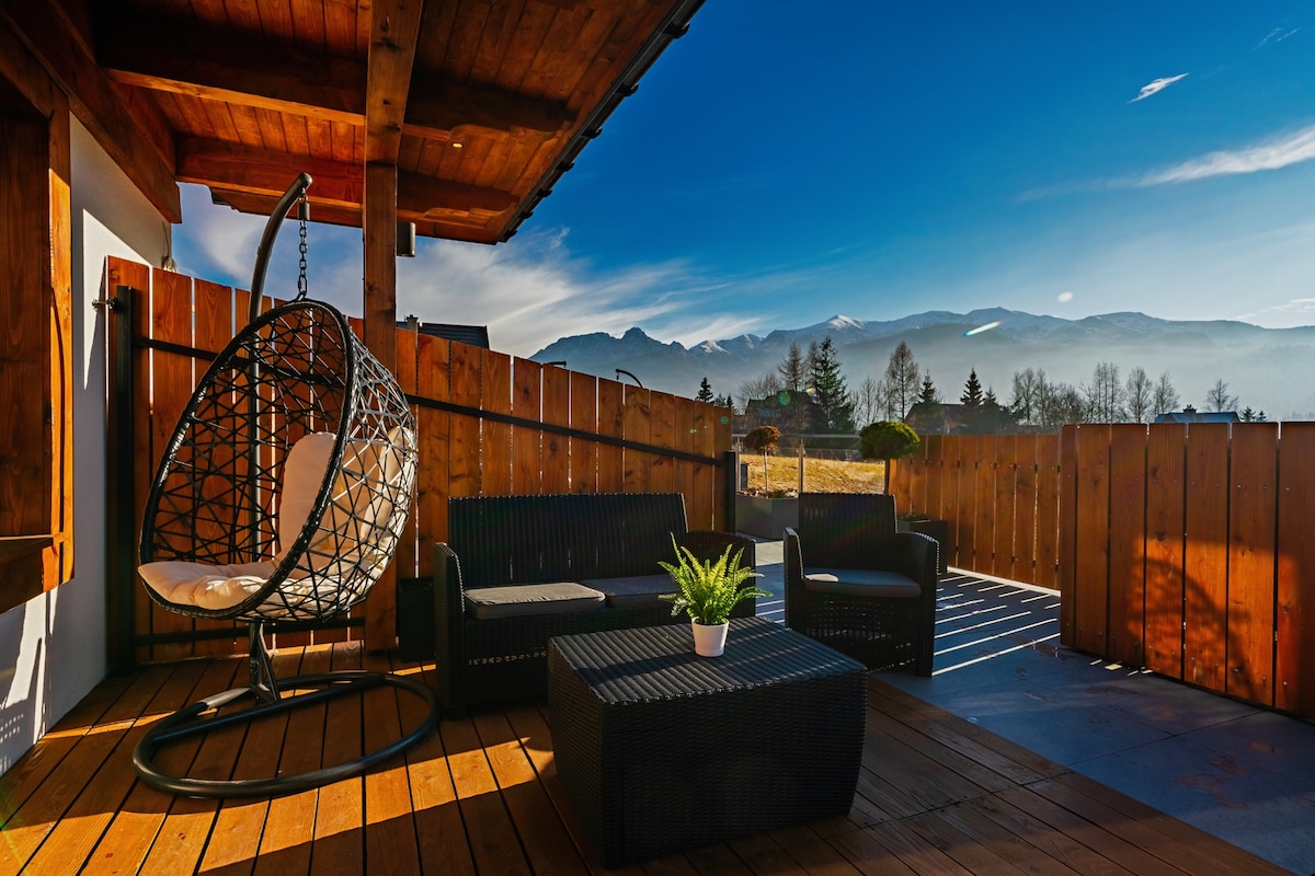 A private terrace features a hanging chair and a black wicker seating arrangement, complemented by a small potted plant. The backdrop showcases a panoramic view of mountains under a clear blue sky, providing a serene outdoor space for relaxation.