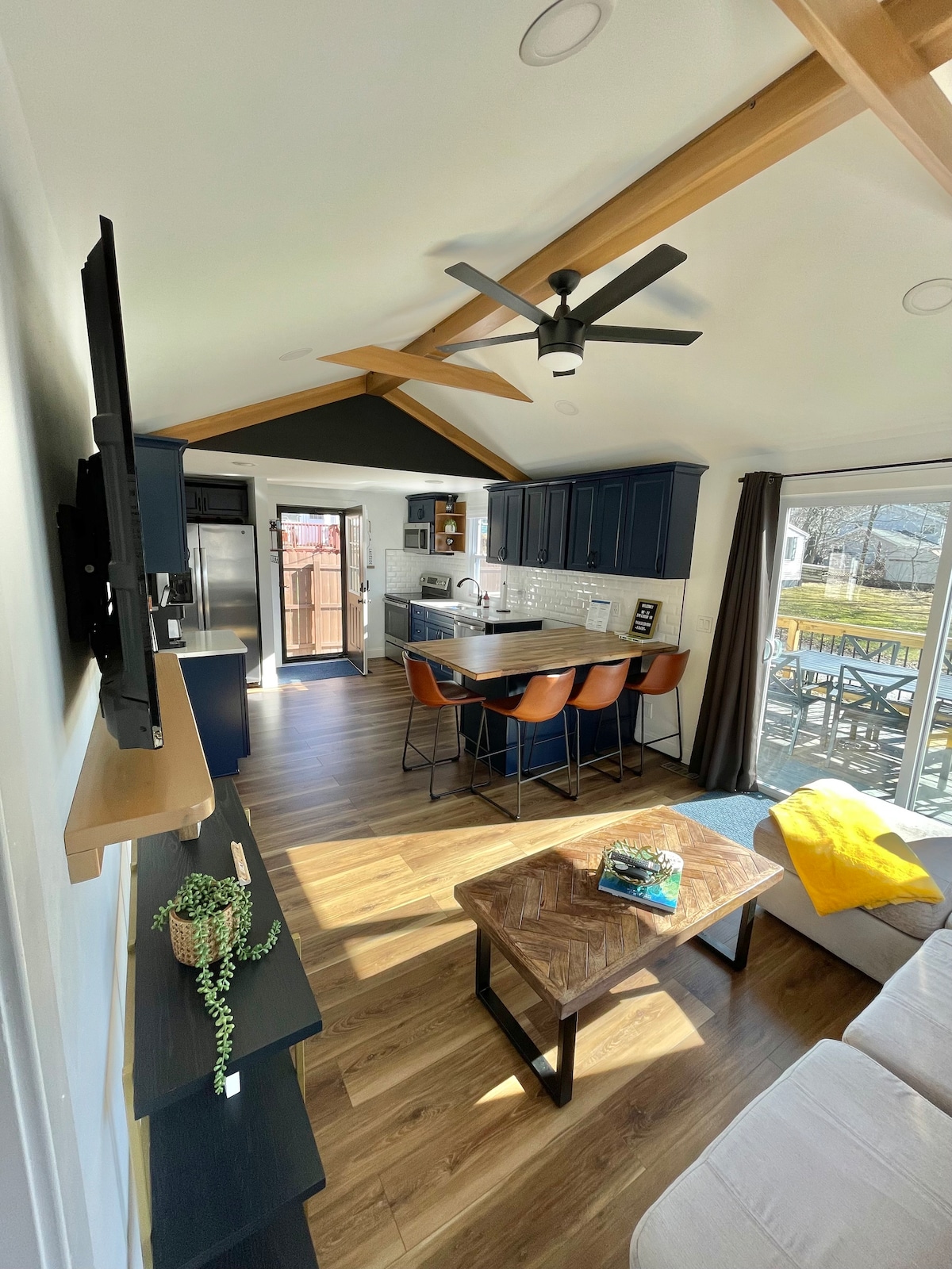 An open and airy living space is highlighted by vaulted ceilings and natural light. The modern kitchen features dark cabinetry and a breakfast bar with four stools. A light-colored couch and wooden coffee table are positioned near a large sliding door, providing access to the outdoor deck.