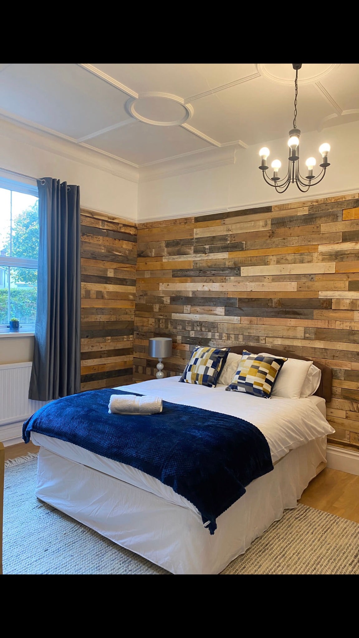 A cozy bedroom features a bed with a white coverlet and navy blue blanket, adorned with two patterned pillows. A wall composed of wooden planks adds texture, while a chandelier hangs overhead. Light filters in through a window dressed with gray curtains.