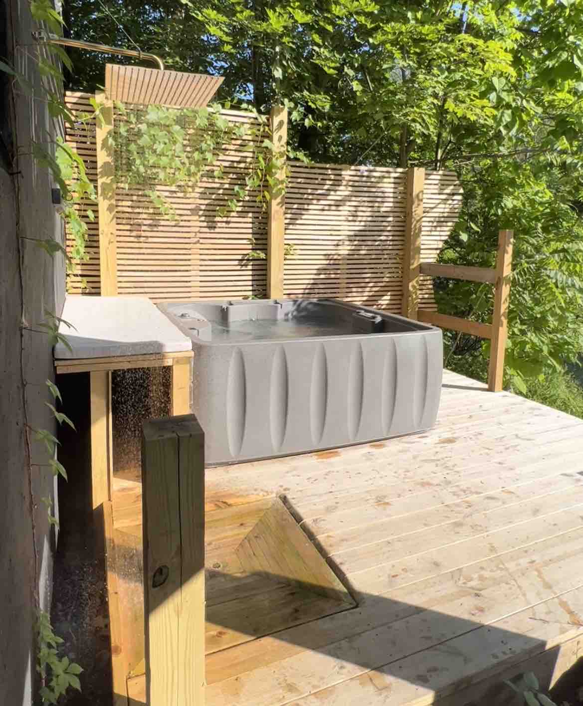 A hot tub is positioned on a wooden deck, surrounded by lush greenery and slatted wooden screens. Water can be seen glistening on the deck, emphasizing a relaxing ambiance. The area offers privacy while inviting soaking experiences in a natural setting.
